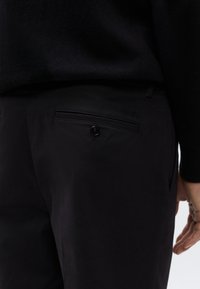 Μαύρο παντελόνι με λεία υφή, που διαθέτει πίσω τσέπη με κούμπωμα κουμπιού και διακριτικό λογότυπο. Απλός σχεδιασμός, εφαρμογή ραμμένη στα μέτρα.