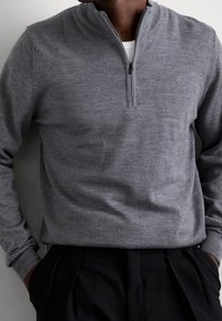 Pull-over gris en tricot avec col à fermeture éclair partielle, poignets côtelés et texture lisse, assorti à un pantalon sombre. Détail discret du logo sur la poitrine.