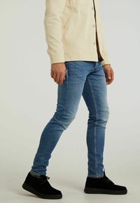 Man som bär en ljusbeige långärmad skjorta, blå smala jeans och svarta skor, stående mot en enkel bakgrund.