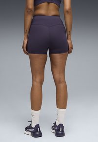 Γυναίκα που στέκεται πλάτη, φοράει σκούρα μωβ αθλητικά σορτς και τοπ, λευκές κάλτσες Puma και μωβ αθλητικά παπούτσια Puma, γκρι φόντο.