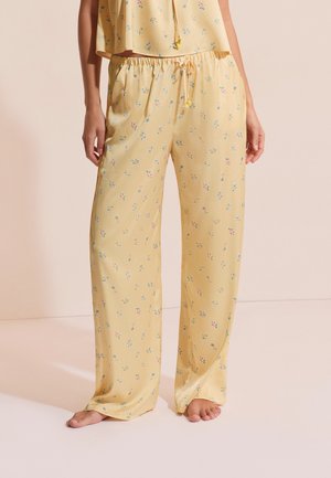 Luźne żółte spodnie do piżamy w drobny kwiatowy wzór, noszone na boso przez osobę stojącą na jasnej podłodze na tle beżowej ściany.