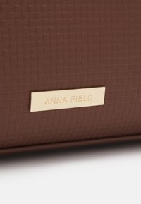 Brązowa teksturowana powierzchnia z prostokątną, złotą metalową płytką wykonaną w technologii grawerowania z nazwą marki "Anna Field."