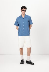 Lyhythihainen sininen chambray-paita, jossa on terävä kaulus, valkoiset shortsit, mustat liukuva kengät ja harmaat aurinkolasit. Minimalistinen muotoilu, siistit linjat.
