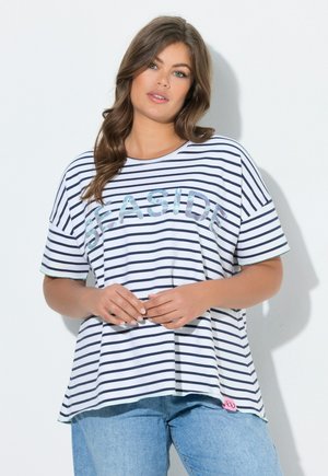 Vrouw met lang bruin haar, die een wit en navy gestreept shirt draagt met de tekst "SEASIDE" en lichtblauwe spijkerbroek, staand tegen een witte muur.