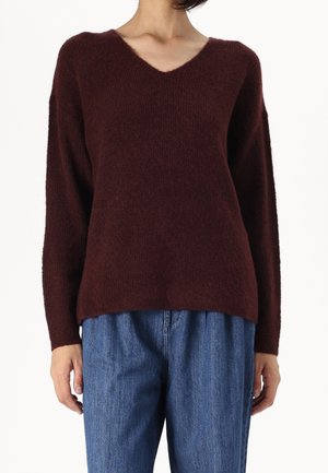 Personne portant un pull à col en V marron foncé en laine pelucheuse avec des manches longues et un jean bleu ample sur fond blanc.