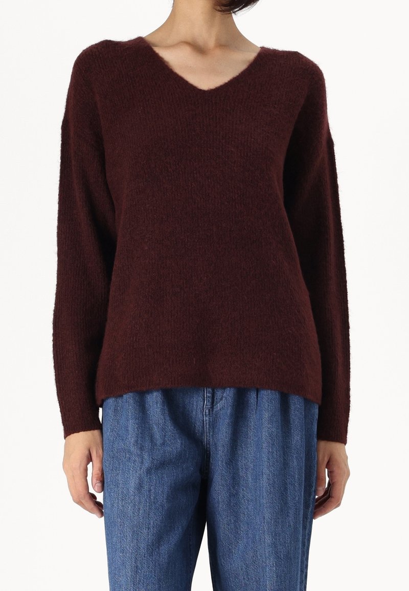 Personne portant un pull à col en V marron foncé en laine pelucheuse avec des manches longues et un jean bleu ample sur fond blanc.