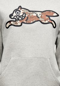 Στυλιζαρισμένη καρτούν αλεπού με κοφτερές οδόντες και καμουφλαρισμένο μοτίβο, που πηδάει πάνω από το στήθος ενός ανοιχτογκρι φούτερ.