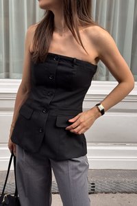 Top nero senza spalline con bottoni e due tasche a pattina, abbinato a pantaloni grigi. Tessuto liscio con vestibilità sartoriale e linee pulite.