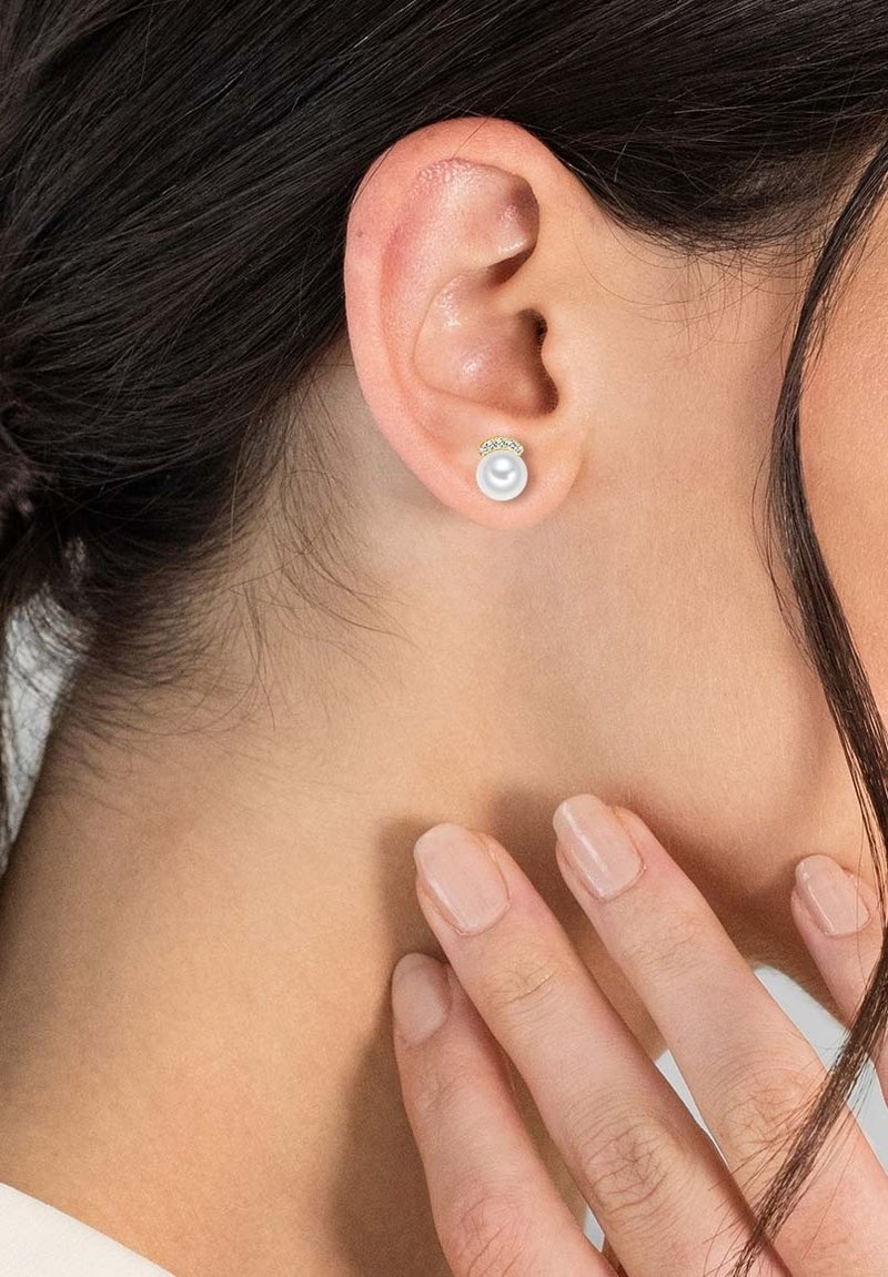 Boucles d'oreilles en perles avec un petit accent décoratif en or. Présentent des perles blanches rondes et lisses, mises en valeur par un teint de peau neutre.