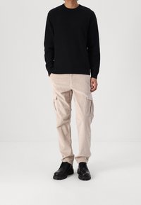 Mann steht mit den Händen in den Taschen, trägt einen schwarzen Pullover, beige Cord-Hose und schwarze Schnürschuhe vor einem weißen Hintergrund.