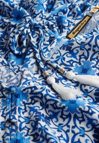 Tela azul con estampado floral, que presenta un fondo blanco. Incluye un cordón con borlas decorativas y acentos metálicos.