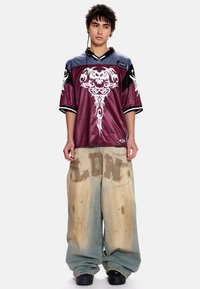 Μακρυμάνικη βυσσινί φανέλα με λευκό γραφικό σχέδιο και μαύρες λεπτομέρειες· φαρδιά ξεθωριασμένα μπλε τζιν με καφέ στάμπα "LDN", φθαρμένη υφή και λεκέδες.