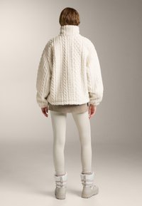 B�ílý oversized svetr s pleteným vzorem a vysokým límcem, texturovaná látka, kleslé ramena, kombinovaný se světle šedými legínami a zateplenými botami.