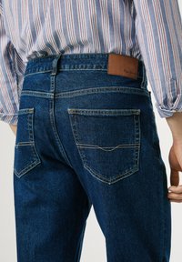 Jeans de mezclilla azul oscuro con una etiqueta de cuero en la parte posterior de la cinturilla, que cuentan con dos bolsillos traseros con costuras en contraste.