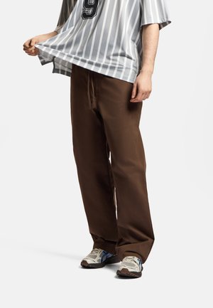 Osoba ubrana w brązowe spodnie o szerokich nogawkach, białe i niebieskie tenisówki oraz szarą koszulę w cienkie paski z numerem 9, trzymającą brzeg koszuli na zewnątrz.