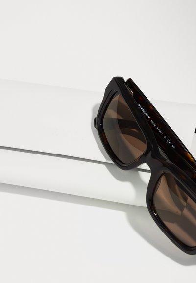 Hnedé korytnačie slnečné okuliare s veľkými obdĺžnikovými rámami, tmavými sklami a výrazným brandingom Burberry na lineárnej časti, položené na bielom povrchu.