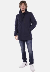 Giacca blu marino in tessuto liscio, con colletto alto, chiusura con zip e tasche laterali, abbinata a jeans blu e sneakers bianche.