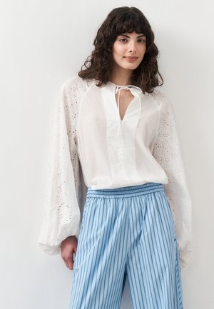 Kobieta z kręconymi ciemnymi włosami nosi białą koronkową bluzkę z bufkami oraz jasnoniebieskie, szerokie spodnie w paski, stojąc na tle białej ściany.