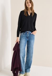 Černé tričko s dlouhým rukávem a výstřihem do V, světlé modré džíny s širokými nohavicemi a texturovaný tmavě fialový svetr v ruce. Neformální outfit s jemnými texturami.