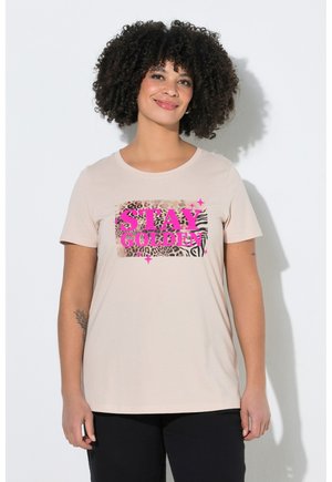 Mujer con cabello rizado que lleva una camiseta beige con texto rosa "STAY GOLDEN" sobre un estampado animal, de pie contra una pared blanca.