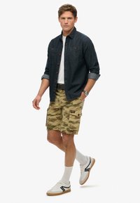 Denim skjorte med knap, hvid T-shirt under, camouflage cargo shorts. Hvide sneakers med sorte detaljer og ankelsokker.
