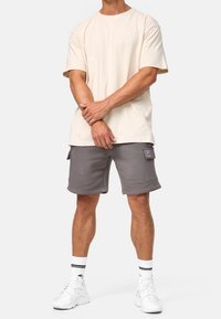 T-shirt en coton beige à manches courtes associé à un short gris avec poches latérales. Baskets blanches avec chaussettes rayées noires et blanches.