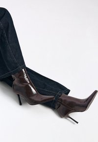 Brune laklæder ankelstøvler med spidse tæer og stiletter. Kombineret med mørk denim bukser, der fremhæver et elegant design og en blank tekstur.