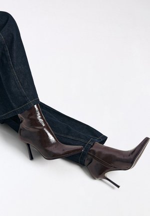 Brune laklæder ankelstøvler med spidse tæer og stiletter. Kombineret med mørk denim bukser, der fremhæver et elegant design og en blank tekstur.