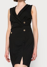 Robe noire sans manches avec un décolleté en V style enveloppant, quatre boutons dorés à l'avant et une fente à la cuisse, portée par une femme aux cheveux courts.