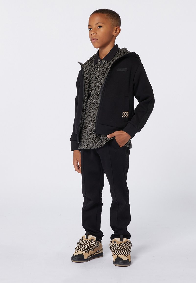 Giacca a zip nera con cappuccio sopra una camicia a fantasia e pantaloni neri. Le calzature presentano una base beige con intricati dettagli in bianco e nero.