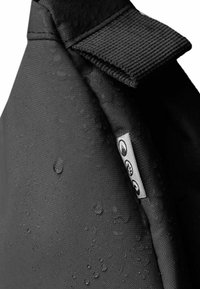 Tessuto impermeabile nero con superficie texture e gocce d'acqua. Presenta una semplice linguetta e un piccolo tag metallico con icone.