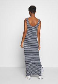 Frau steht mit dem Rücken zu uns und trägt ein ärmelloses-maxikleid mit navy- und weißen Streifen sowie einen seitlichen Schlitz und weiße Sneakers.