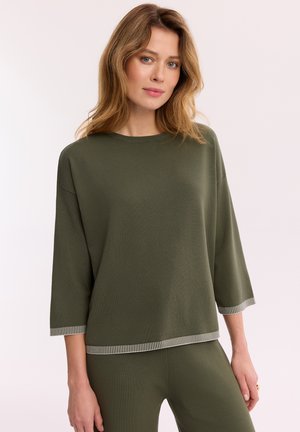 Kobieta ubrana w luźną oliwkowozieloną bluzkę i pasujące spodnie z białym paskowanym wykończeniem na rękawach i dole, na jasnym jednolitym tle.