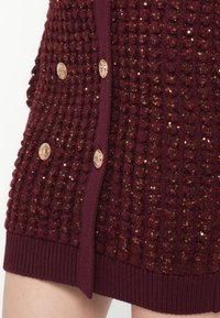 Maglione bordeaux con un design a maglia texturizzata e a pallini, bottoni dorati e orlo a costine. Presenta una leggera lucentezza data da paillettes.
