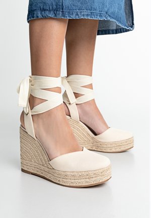 Espadrilles à talon compensé beige avec une semelle en jute tissée, présentant un dessus en toile et des nouages en tissu entrecroisés au niveau de la cheville. Fini lisse, design à bout ouvert.