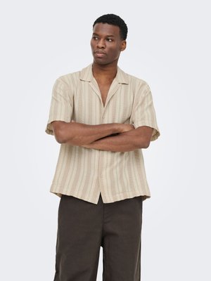Mann steht mit verschränkten Armen, trägt ein beige gestreiftes kurzärmeliges Hemd mit Knöpfen und dunkelbraune Hose, blickt leicht zur Seite.