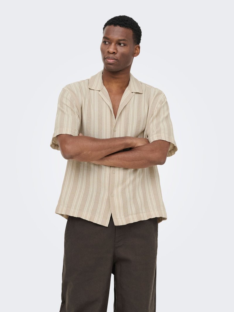 Homme debout, les bras croisés, portant une chemise beige à manches courtes rayée et boutonnée, ainsi qu’un pantalon marron foncé, regardant légèrement sur le côté.