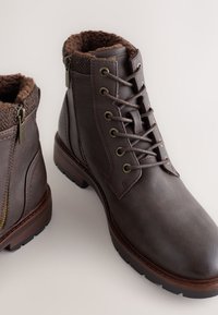 Bottes en cuir marron montant à la cheville, dotées d'une doublure en fourrure marron douce et texturée, de ferrures métalliques et d'une semelle robuste avec des accents de motifs.