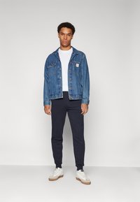 Giacca di jeans blu con due tasche sul petto, t-shirt bianca, pantaloni della tuta blu navy e sneakers bianche con dettagli beige. Tessuti lisci in tutto l'outfit.