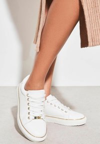 Adidași albi cu design cu șireturi, accente aurii, vârf rotund și talpă căptușită. Partea superioară are o textură netedă și detalii minime.