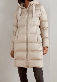 Abrigo acolchado beige con capucha, cierre de cremallera, dos bolsillos delanteros y diseño acolchado. Textura suave con correas de acento.