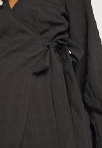 Vestido preto envolvente feito de tecido texturizado. Apresenta um detalhe de laço na cintura e mangas longas e fluidas com uma superfície enrugada.