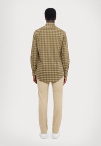 Man som står med ryggen mot kameran och har på sig en långärmad grön och beige rutig skjorta, beige byxor och vita sneakers mot en enfärgad bakgrund.