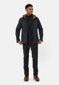 Męska ciemnoniebieska kurtka softshell z jasnobrązowym podszyciem, kieszeniami na zamek i eleganckim krojem, zestawiona z czarnymi spodniami i brązowymi butami.