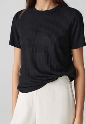 Kobieta ubrana w czarny prążkowany top z krótkim rękawem, lekko wpuszczony do białych spodni z wysokim stanem na jednolitym tle.