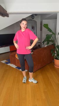 Femme portant un haut rose vif et un legging noir, posant les mains sur les hanches dans un salon avec un sol en bois et des plantes.