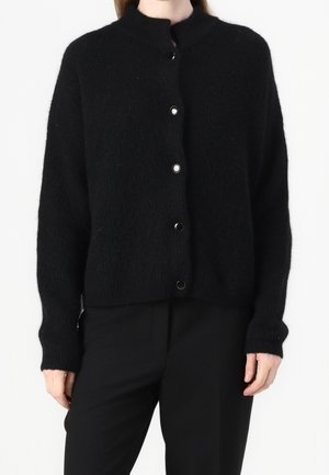 Femme portant un cardigan en maille duveteuse noire avec un col rond et cinq boutons-pression visibles sur le devant, associé à un pantalon noir.