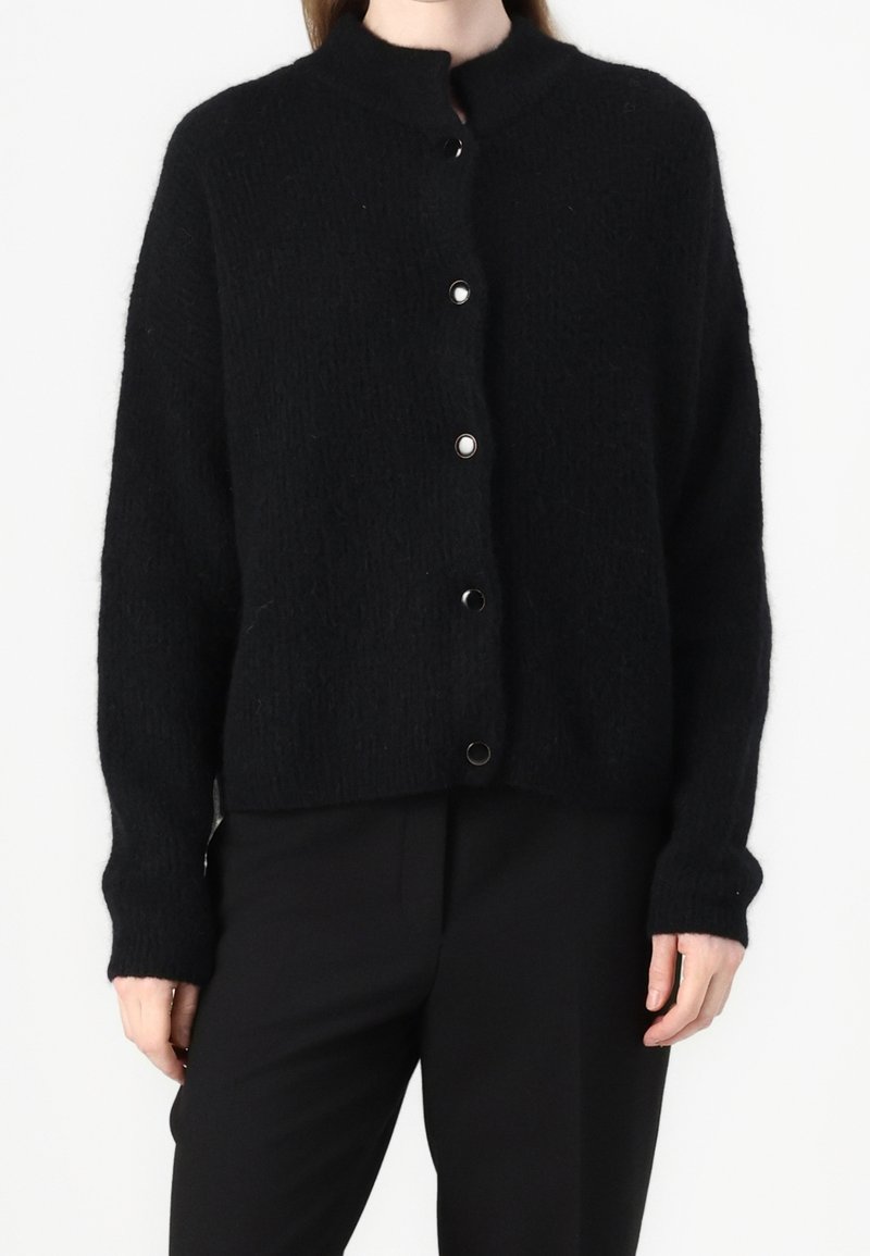 Femme portant un cardigan en maille duveteuse noire avec un col rond et cinq boutons-pression visibles sur le devant, associé à un pantalon noir.