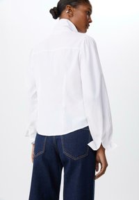 Blouse blanche avec un col montant, des manches bouffantes longues et des poignets boutonnés. Associée à un jean large bleu foncé avec des coutures visibles.