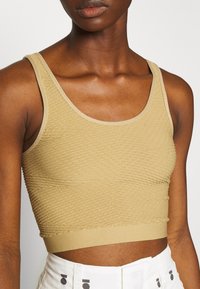 Zkrácený top bez rukávů ve světle béžové barvě s texturovaným povrchem a kulatým výstřihem. Má přiléhavý lem a široké ramínka.